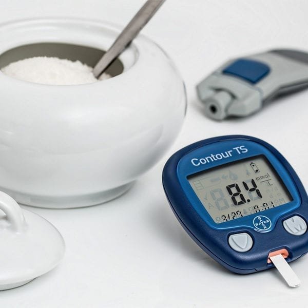 A glucose meter displaying a reading of 8.4 mmol/L, a lancing device, and a bowl of white sugar with a spoon on a white surface.